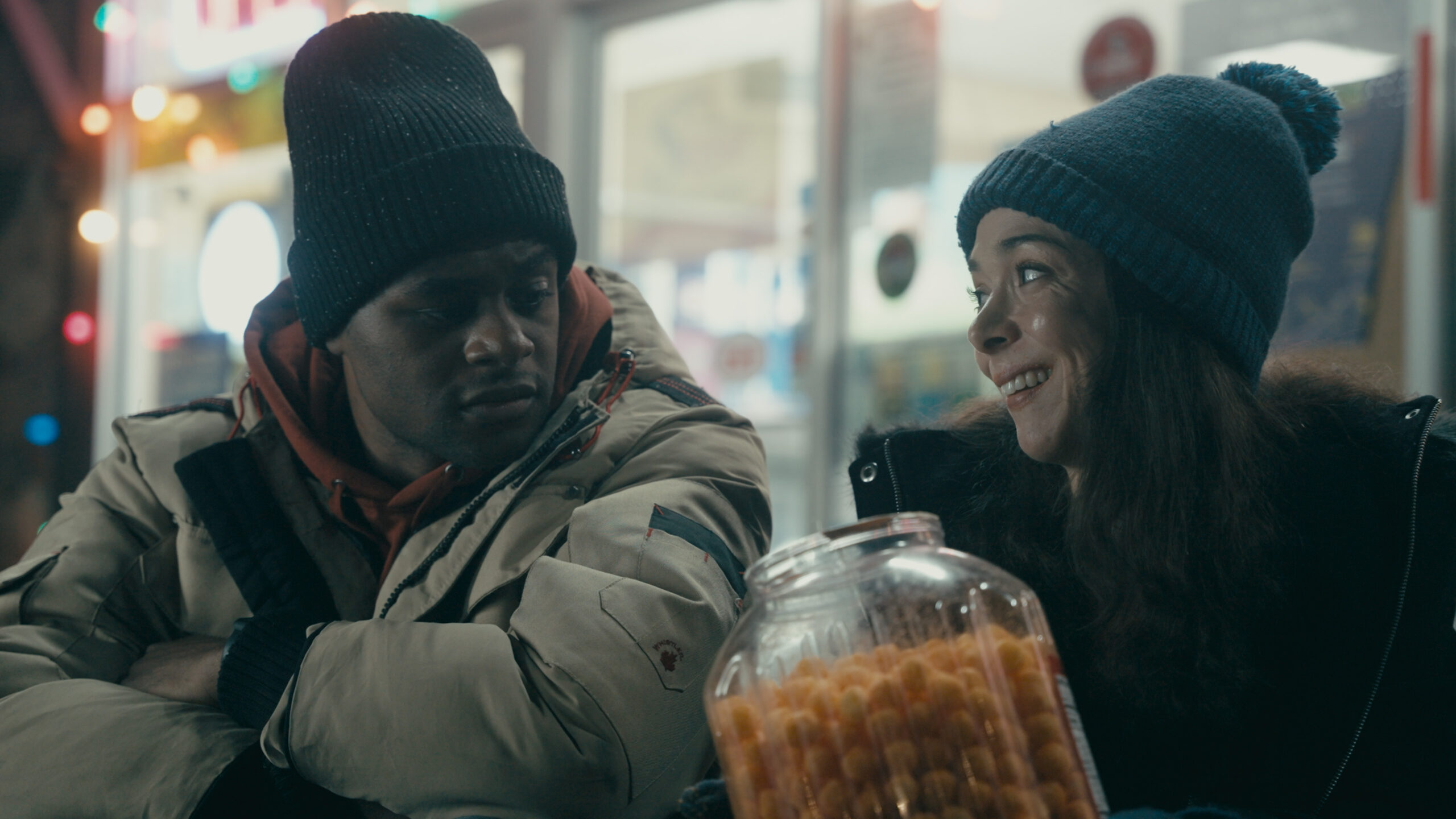 two people dressed in winter coats and hats share snacks outside